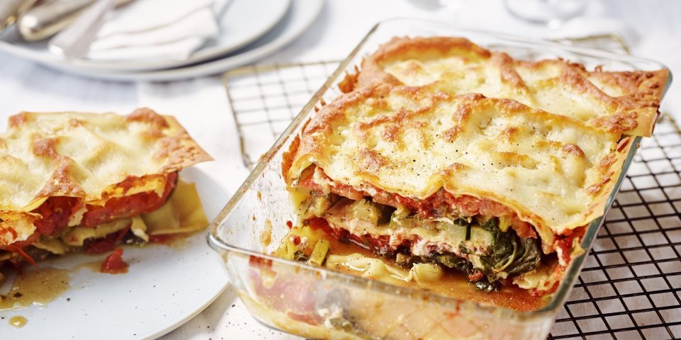 De lasagne staat op tafel in een glazen doorzichtige ovenschotel. Hij is aangesneden zodat je mooi de laagjes ziet van pasta, groenten en saus. De portie die is uitgeschept ligt op een bordje er naast.