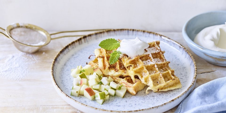 Wafeltjes van verloren brood liggen op een licht grijs bordje. De stukjes fruit van appel en peer liggen onder de wafeltjes. We gaven ook een toefje slagroom op de wafels voor extra smaak. Er ligt een blauwe servette en klein zeefje met bloemsuiker langs het bord.