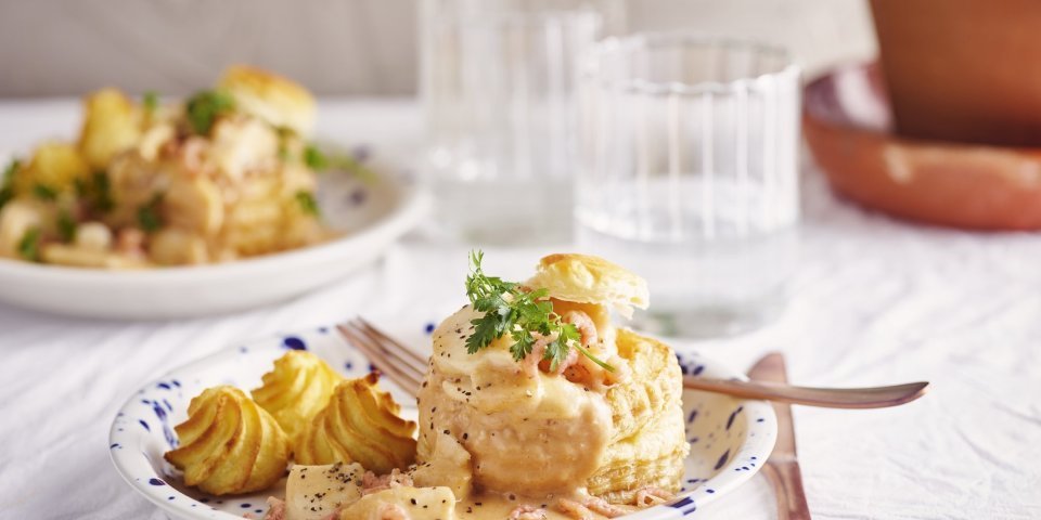 Twee borden staan klaar om van te smullen. Vol-au-vent van vis in een bladerdeegpasteitje met pommes duchesse. Er staat een glaasje water naast het bord.