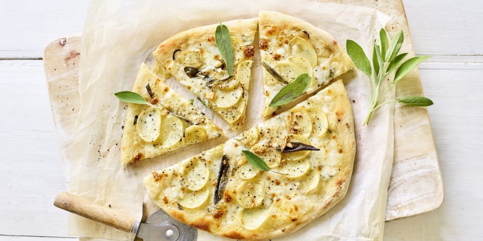 Een pizza belegt met mozzarella, plakjes aardappel en salie ligt op een wit boterpapiertje. Er ligt een pizzasnijder langs de pizza en nog enkele verse blaadjes salie. 