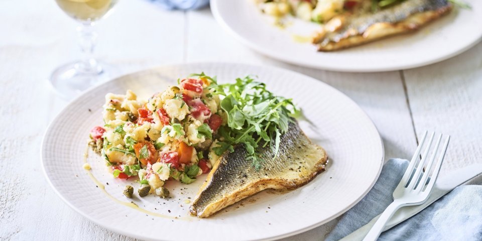 Op tafel staan twee borden met het gerechtje op. Het is een aardappelpuree met stukjes paprika, kappertjes en rucolasla. En een krokant gebakken zeebaarsfilet. Er staat ook een glaasje witte wijn langs. 