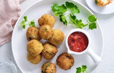 Heerlijke krokante balletjes liggen op een wit bord gepresenteerd. Hier is een klein potje met ketchup bijgeplaatst en enkele blaadjes peterselie voor extra groen. 