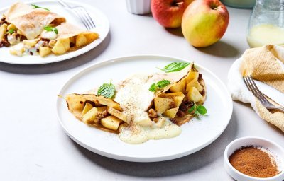 Op tafel staan twee borden met elk hierop een dichtgevouwen pannenkoek, die gevuld is met stukjes appel en rozijnen. Wij serveren hem met vanillesaus en verse blaadjes munt. Er liggen ook nog enkele appelen in de achtergrond en een restje vanillesaus.