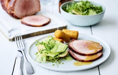 Op een houten witte tafel staat een portie klaar. Een wit bord gevuld met twee sneetjes van het varkensgebraad, kroketten en de salade van komkommer met appel.