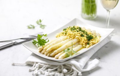 Op een rechthoekig wit bord liggen gekookte asperges, afgewerkt met geplette gekookte eieren, peterselie en gesmolten boter. Klaar om aan te vallen. 