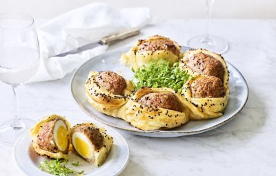 Op tafel staat een bord waarop een bladerdeegkrans ligt. Er zitten grote gehaktballen tussen gevluchten met hierin een gekookt eitje. Het bladerdeeg is afgewerkt met zwarte sesamzaadjes wat zorgt voor een leuke spikkeling.