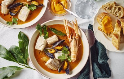 Een heerlijk kleurrijk bordje bouillabaisse staat op tafel. Wij maakten hem met topproducten van hier. Er ligt een donker groene linnen servette langs en bordje stukjes brood en verse rouille.