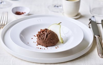 In een diep wit bord ligt een bolletje chocomousse met hierop de crumble van spek en een plakje peer. Bovenaan zie je een heerlijke kopje kofie staan. Perfect dessert om het feestmenu in glans af te sluiten.