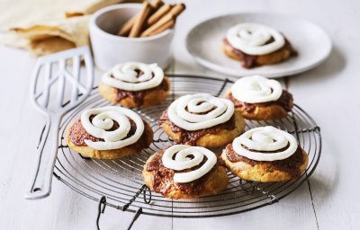 Op een taartrooster liggen 6 koekjes af te koelen. Het zijn koekjes van cinnamon rolls, een nieuwe versie om te genieten van deze heerlijke en kruidige zoetigheid. Op de koekjes is frosting gespoten op basis van roomkaas. 