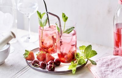 Twee cocktails staan op tafel, ze hebben een mooie roodroze kleur. Onderaan het glas ligt het smached fruit, met daarna de vodka en roze tonic. Er liggen nog enkele aardbeien en kersen langs. 