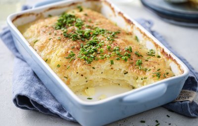 De ovenschotel met gratin staat op tafel, hij heeft een lichtblauwe kleur. De aardappelschijfjes zijn lekker crispy gebakken, doordat we ze in de oven klaarmaken met minder room. De ovenschotel is afgewerkt met verse fijngehakte bieslook.