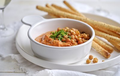 Een wit porseleine potje is gevuld met een oranjekleurige tapenade. Het is er eentje van zoete puntpaprika met hierop geroosterde kikkererwten. Er liggen broodstengels langs om te dippen. 