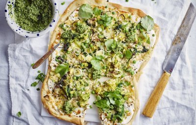 Op een witte snijplank ligt de rechthoekige flammkuchen. Hij heeft prachtige groene tinten vavn de erwtjes, prei en blaadjes waterkers. Er ligt een groot mes langs om aan te snijden een potje met extra pesto van waterkers.