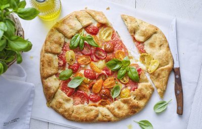 De galette van tomaten ligt op de tafel op een wit boterpapiertje. Hij is mooi gebakken en er is een stukje van gesneden. Er staat een basilicumplantje langs, alsook een flesje gouden olijfolie. 