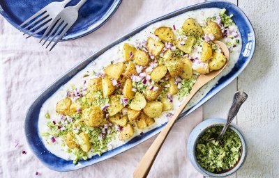 Een langwerpig blauw bord staat op tafel. Hierop is yoghurt op geschept met krokante aardappelen, stukjes rode ui en geraspte broccoli. 