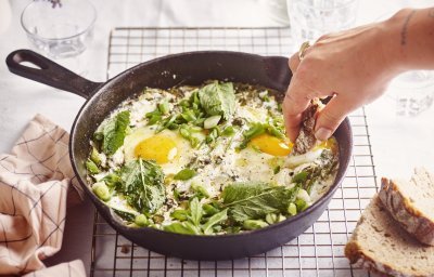 Op een rooster staat een zwarte gietijzeren pan met grieks getint gerechtje. Groene shakshuka met gebakken yoghurt. Er is een hand met een stukje brood in beeld dat er een dipje van zal nemen. 