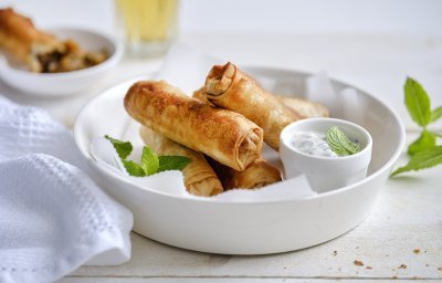 Meerdere rolletjes liggen te pronken in een wit serveerschaaltje. De rolletjes zien er heerlijk krokante gebakken uit en worden geserveerd met een dip van yoghurt en munt in een ander wit klein potje. Er ligt ook nog een takje munt bij voor een vleugje extra groen.