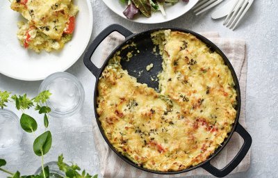 Een taart van aardappelpuree en paprika, staat op het midden van de tafel. Hij zit in een zwarte schotel en heeft een mooi kaaskorstje meegekregen uit de oven. Er is al 1 portie uitgeschept, en er ligt ook bestek op tafel, sla en verse kruiden. 