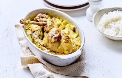 Op tafel staat een witte ovale ovenschotel met hierin de gele currysaus met stukjes appel en ananas. Hier bovenop liggen de bouten te blinken. De ovenschotel staat op een beige linnen handdoek met rijst, borden en een witte pepermolen langs.