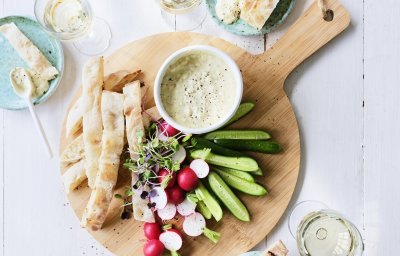 Op tafel staat een ronde houten snijplank met hierop een wit potje gevuld met onze pikante dip van yoghurt en witte bonen. Je kan de yoghurtdip gebruiken met stukjes snackkomkommer, brood en radijsjes in ons geval. Rond de plank staan enkele glazen witte wijn en helderblauwe broodbordjes. 