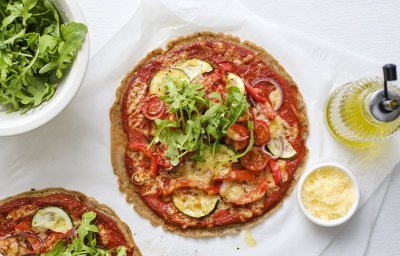 Op tafel staan twee klaargemaakte pizza's op een wit bakpapiertje. Hij is afgewerkt met verse rucola en wat olijfolie.