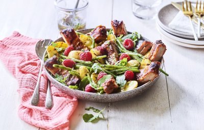 Een grote serveerschaal is gevuld met de aardappelsalade. Je ziet de framboosjes, boontjes en ribbetjes in de salade zitten. Er staat een torentje bordjes klaar met bestek om de salade te verdelen en op te eten. 