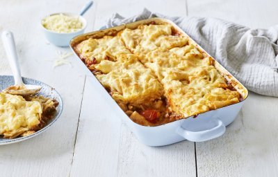 Een lichtblauwe ovenschotel staat op een houten witte tafel. De ovenschotel is gevuld met een mac & cheese én een tomatensaus, waardoor hij eerder weg heeft van een lasagne. Er is al 1 portie uitgeschept en wil al beginnen aan de volgende. Laat het niet koud worden. 