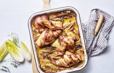 Op een grote houten snijplank staat de ovenschaal met de tartiflette en stukken konijn. Het konijn in omwikkeld met sneetjes spek, waardoor het nog iets extra heeft. Er ligt witloof langs en verse tijm.