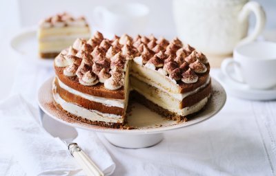 Op tafel staat een prachtige taart, het is een twist op de klassieke tiramisu. Hij staat op een witte taartschaal, en is afgewerkt met toefjes mascarpone en cacaopoeder. In de achtergrond staat een koffiekan en tasjes koffie.