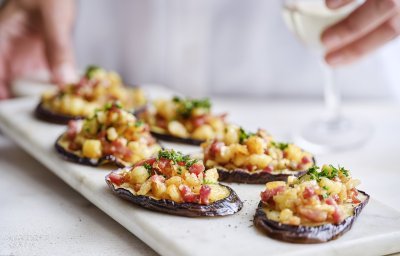 Op een marmere serveerschaal liggen enkele aubergineplakjes. Deze dienen als toastje en zijn belegd met stukjes Berloumi en spek. 