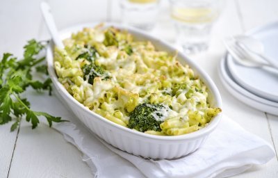 Een grote ovenschaal staat op een houte witte tafel. Hij staat op een wit lapje stof. De ovenschotel is gevuld met macaroni, broccolie en witte bloemkoolsaus. Er staan enkele bordjes langs met wit bestek om direct aan te vallen. 