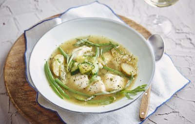 Een heerlijke maaltijdsoep is geserveerd in een wit soepbord. Deze staat op tafel, op een houte ronde snijplank, op een witte servette met blauwe rand. Het bord is gevuld met de doorschijnende bouillon met hierin de stukjes groenten en vis. Echt een feestsoepje. 