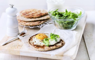 Het yoghurtbrood ligt op een houten snijplank. Hierop liggen meerdere ingrediënten om aan de slag mee te gaan, denk maar aan rucola, peper, extra yoghurt. 