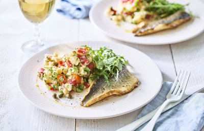 Op tafel staan twee borden met het gerechtje op. Het is een aardappelpuree met stukjes paprika, kappertjes en rucolasla. En een krokant gebakken zeebaarsfilet. Er staat ook een glaasje witte wijn langs. 