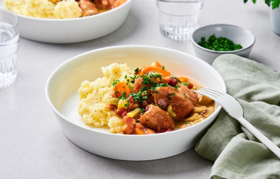 Op tafel staan twee uitgeschepte porties van het hoofdgerecht. Een schep heerlijke puree met kalfsfricassee. In de fricassee zitten spekblokjes, selder en wortelstukjes. Het gerechtje is afgewerkt met verse peterselie. Er ligt een licht groene servette langs en wit bestek.