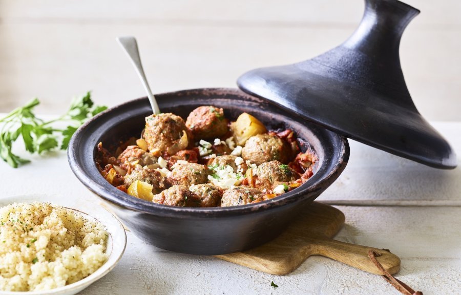 Zwartkleurige tajine met gehaktballetjes van lamsvlees, afgewerkt met verbrokkelde feta. Er staat een portie couscous naast de tajine. 