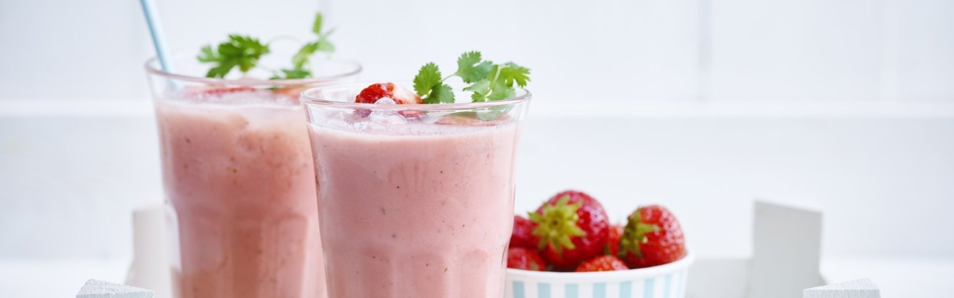 2 smoothies van aardbeien en melk. Ze staan in een houten bakje met een portie verse aardbeien er naast. De smoothies zijn afgewerkt met een takje koriander en kartonnen licht blauw rietje