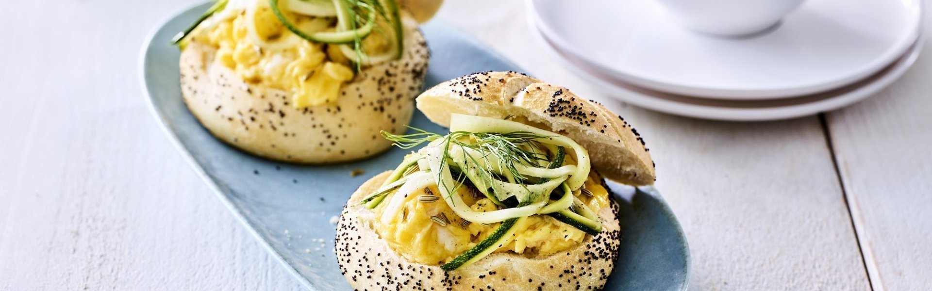 Twee pistolets liggen op een grijsblauw bordje. De pistolets zijn opengesneden en gevuld met roerei en courgetteslingers. 