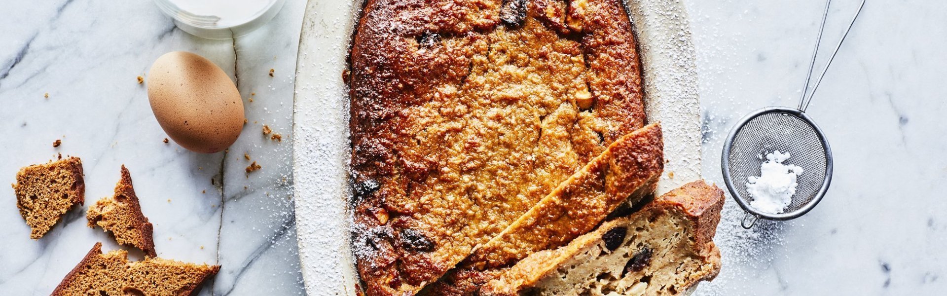 Een heerlijke homemade broodpudding staat op een marmer aanrecht in een witte ovenschaal. Hij is al aangesneden en een stukje uit de vorm genomen. Om de foto meer boddy te geven ligt er een eitje, flesje melk, stukje peperkoek en bloemsuiker naast.