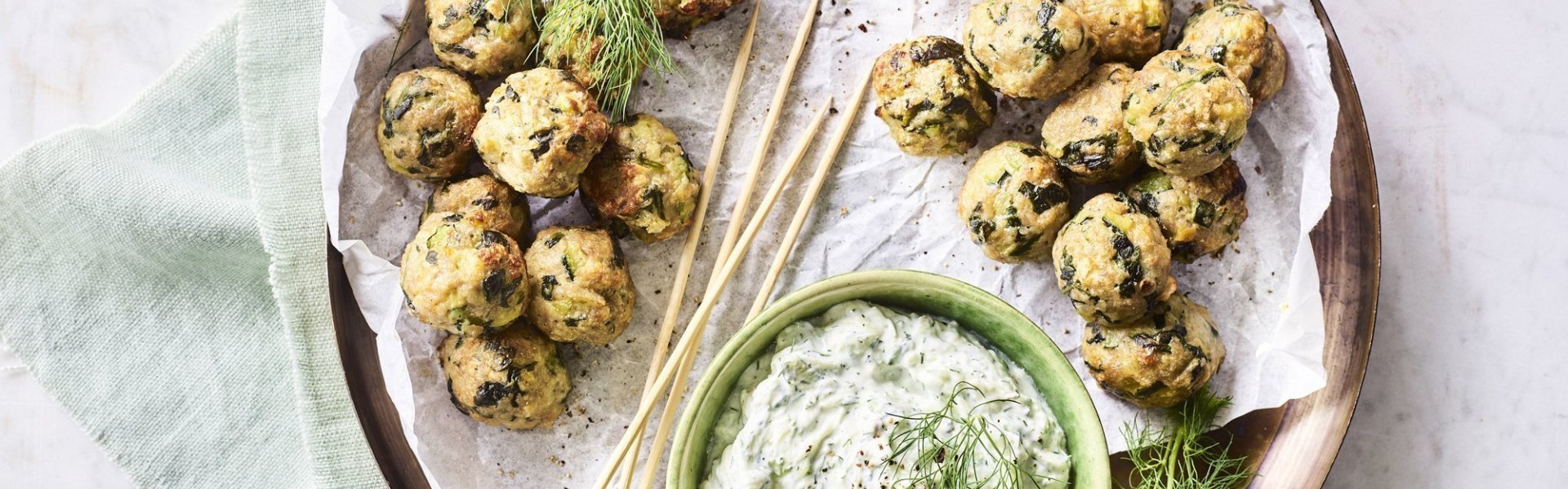 Op het aanrecht staat een houten ronde serveerschaal met een wit boterpapiertje op. Hierop liggen kleine gehaktballetjes met groene stukjes in van de courgette en spinazie. De dip op basis van tzatziki staat ook op de schaal. Het gerecht is afgewerkt met enkele takjes dille. 