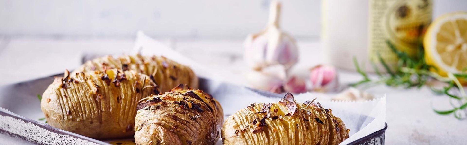 Klaargemaakte hasselback potatoes met pittige kruiden. Ze liggen op een grijze ovenschaal op de keukentafel. Achteraan het beeld zie je hete pepers, look, rozemarijn en een halve citroen liggen.