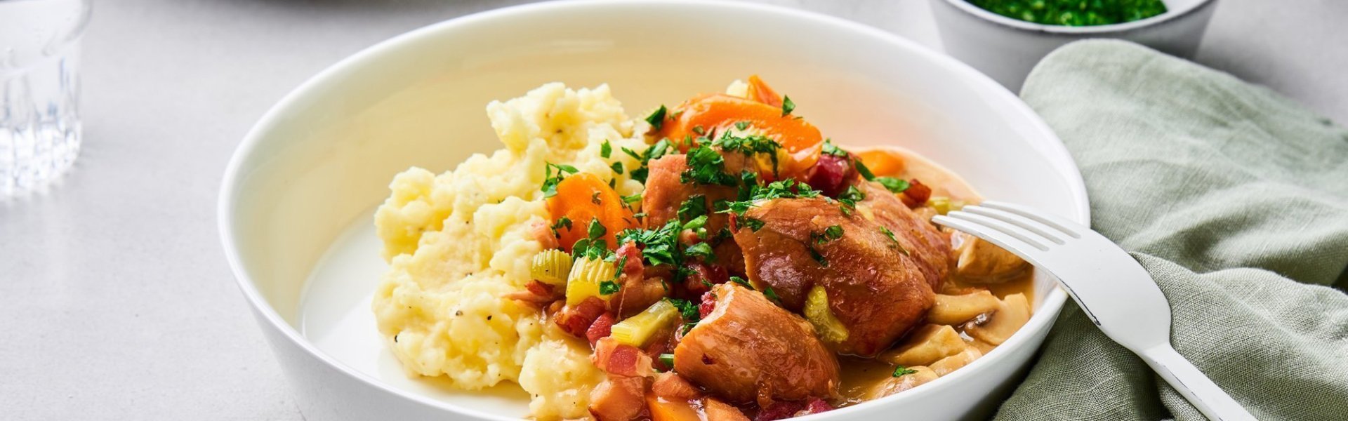 Op tafel staan twee uitgeschepte porties van het hoofdgerecht. Een schep heerlijke puree met kalfsfricassee. In de fricassee zitten spekblokjes, selder en wortelstukjes. Het gerechtje is afgewerkt met verse peterselie. Er ligt een licht groene servette langs en wit bestek.