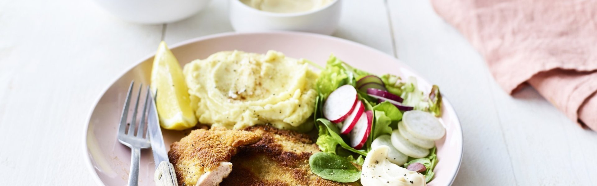 Een homemade shnitzel ligt op een pastelroze bord, samen met aardappelpuree, verse sla en zelfgemaakte mayonaise. Er is nog wat extra salade in een wit potje en mayonaise voor extra lekkers op te scheppen. 