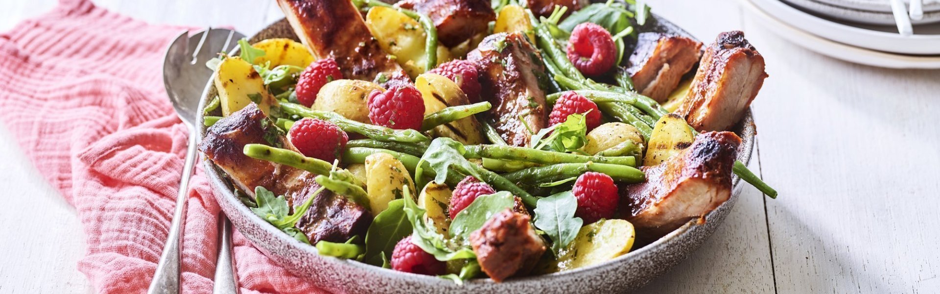 Een grote serveerschaal is gevuld met de aardappelsalade. Je ziet de framboosjes, boontjes en ribbetjes in de salade zitten. Er staat een torentje bordjes klaar met bestek om de salade te verdelen en op te eten. 