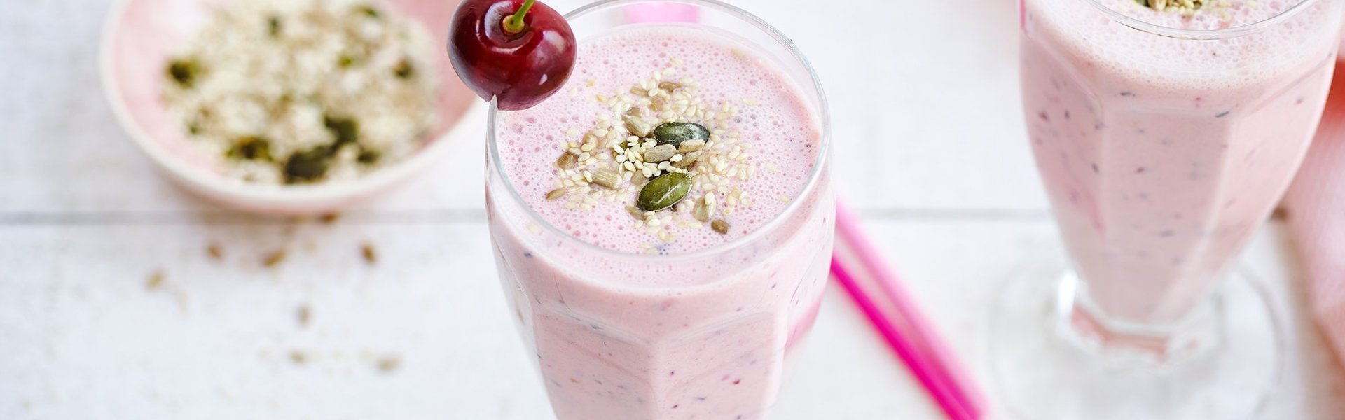 Twee rooskleurige smoothies op een witte houten tafel. De smoothie is afgewerkt met een kers en enkele pitjes en zaadjes. Een snel en makkelijk drankje met feestallures! 