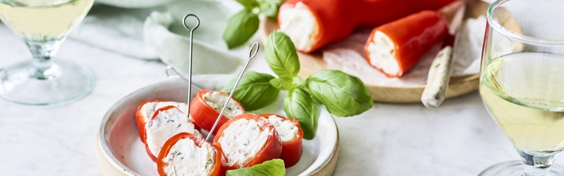 Een opgevulde puntpaprika met roomkaas en kruiden ligt op een houten snijplankje. Er werden rolletjes van gesneden en vastgeprikt met een stokje om het aperitief in stijl te starten. Voor de garnering en de kleur voegde we nog enkele verse blaadjes basilicum toe.