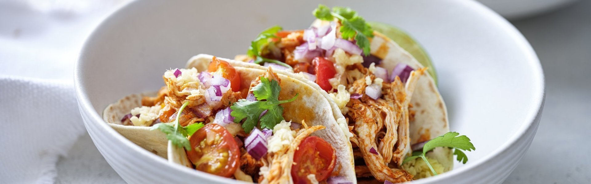 In een diep wit bord liggen twee gevulde taco's met pulled chicken, afgewerkt met tomaat, rode ui en geraspte Passendael kaas. Om wat extra kleur te geven liggen er enkele blaadjes koriander op. Wij serveren ze met frisgekoeld biertje, smakelijk!