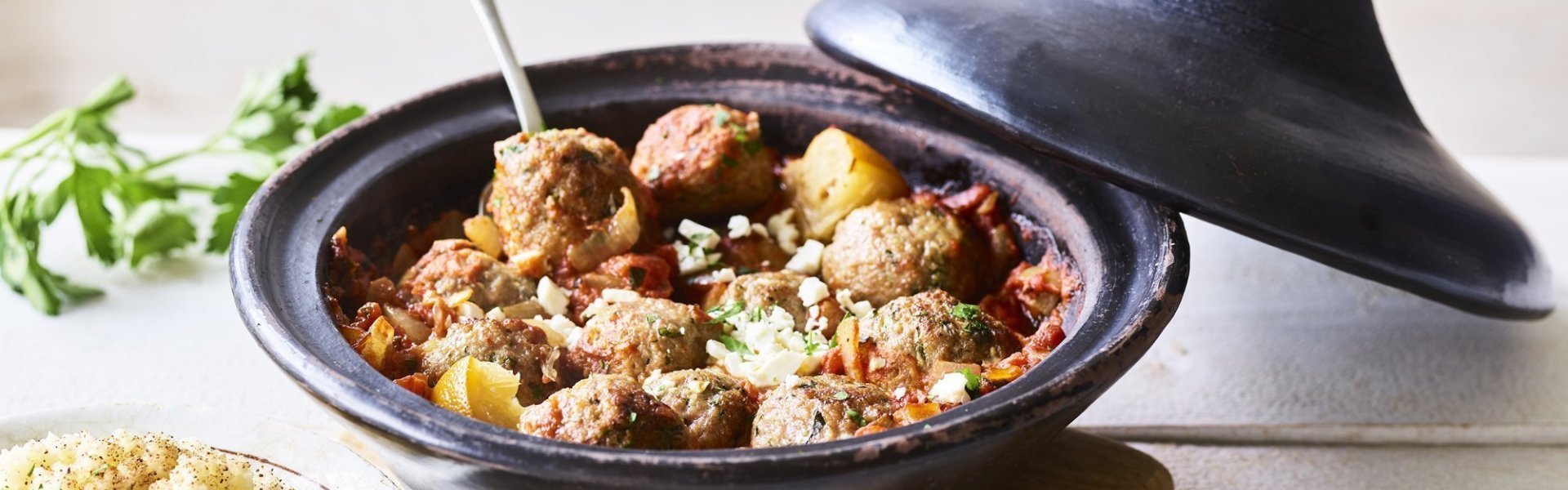 Zwartkleurige tajine met gehaktballetjes van lamsvlees, afgewerkt met verbrokkelde feta. Er staat een portie couscous naast de tajine. 