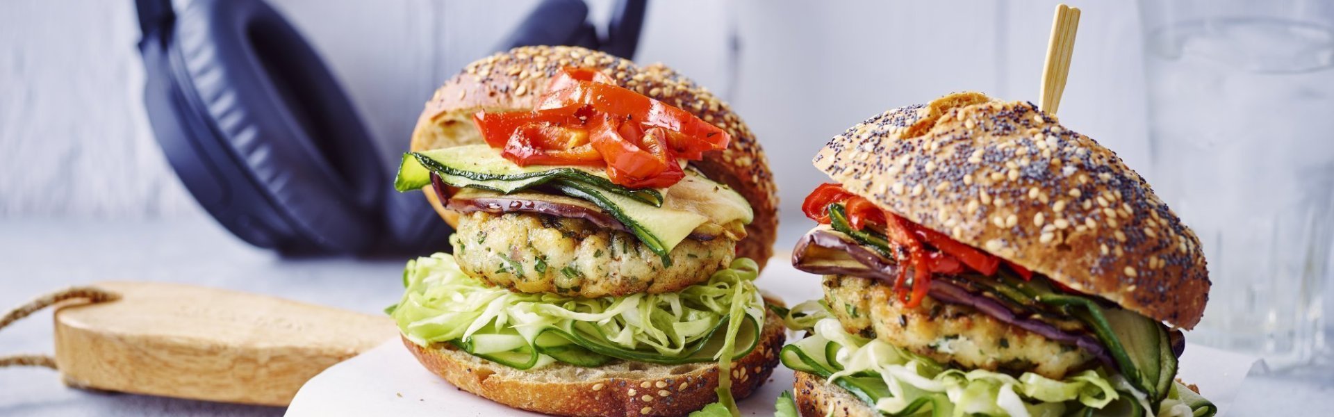 Een visburger tussen een bruine pistolet met veel groenten. Hij is opgebouwd in verschillende secties, onderaan spitskool daarna komkommerplakjes, de visburger, warme courgette, aubergine en puntpaprika en we sluiten af met het hoedje van de pistolet. Op de foto zie je ook een koptelefoon liggen op het aanrecht.