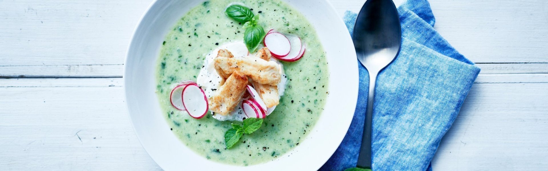 Wit diep bord met gazpacho van komkommer, tongscharreepjes, radijsjes en basilicum. Rechts naast het bord ligt een lepel op een blauw servette.